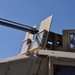 The image shows a close-up, low-angle view of a military vehicle's weapon turret system mounted on what appears to be a tan/desert-colored armored vehicle. A multi-barrel rotary cannon (consistent with a M134 Minigun or similar Gatling-style weapon) is visible along with an ammunition belt feed. A soldier in military gear and eye protection is visible through a side viewport of the vehicle.