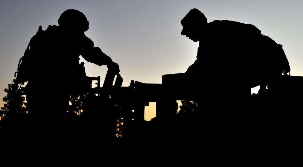 A dramatic silhouette photograph taken at dusk or dawn showing two military personnel working on or around what appears to be a military vehicle. The figures are completely backlit against a gradient sky transitioning from orange near the horizon to blue-gray above. The image captures a moment of operational activity with strong compositional contrast between the dark foreground subjects and the illuminated sky.