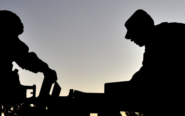 A dramatic silhouette photograph taken at dusk or dawn showing two military personnel working on or around what appears to be a military vehicle. The figures are completely backlit against a gradient sky transitioning from orange near the horizon to blue-gray above. The image captures a moment of operational activity with strong compositional contrast between the dark foreground subjects and the illuminated sky.