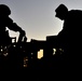 A dramatic silhouette photograph taken at dusk or dawn showing two military personnel working on or around what appears to be a military vehicle. The figures are completely backlit against a gradient sky transitioning from orange near the horizon to blue-gray above. The image captures a moment of operational activity with strong compositional contrast between the dark foreground subjects and the illuminated sky.