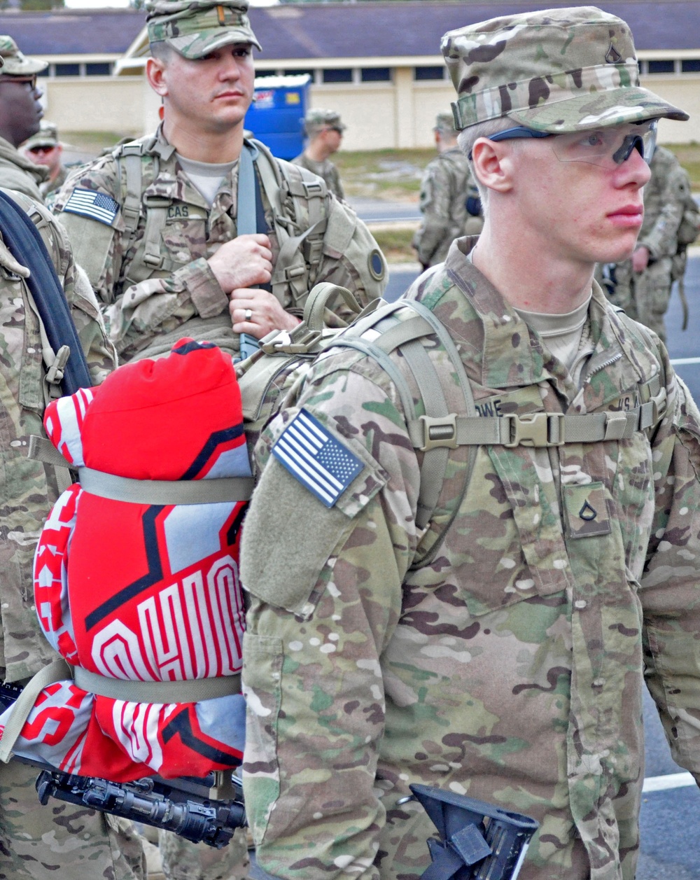 A group of U.S. Army soldiers in Operational Camouflage Pattern (OCP) uniforms are assembled in formation, likely at a military installation. The foreground soldier carries a rucksack with an Ohio State Buckeyes blanket or clothing item strapped to it. Soldiers are equipped with rifles, tactical gear, and standard-issue equipment.