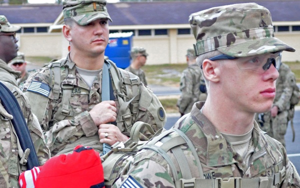 A group of U.S. Army soldiers in Operational Camouflage Pattern (OCP) uniforms are assembled in formation, likely at a military installation. The foreground soldier carries a rucksack with an Ohio State Buckeyes blanket or clothing item strapped to it. Soldiers are equipped with rifles, tactical gear, and standard-issue equipment.