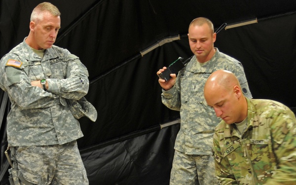 Three U.S. Army soldiers are working with an unmanned aerial vehicle (UAV/drone) in what appears to be an indoor military facility or tent. One soldier in Operational Camouflage Pattern (OCP) uniform is actively working on the UAV, while a second soldier in Army Combat Uniform (ACU) holds a handheld device, and a senior soldier observes with arms crossed. The scene appears to be a maintenance, inspection, or demonstration event.