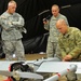Three U.S. Army soldiers are working with an unmanned aerial vehicle (UAV/drone) in what appears to be an indoor military facility or tent. One soldier in Operational Camouflage Pattern (OCP) uniform is actively working on the UAV, while a second soldier in Army Combat Uniform (ACU) holds a handheld device, and a senior soldier observes with arms crossed. The scene appears to be a maintenance, inspection, or demonstration event.