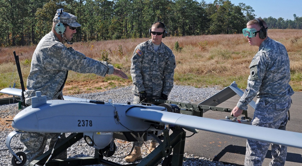 Three U.S. Army soldiers in ACU (Army Combat Uniform) camouflage are preparing or inspecting a small unmanned aerial vehicle (UAV) marked with the number '2378'. The aircraft appears to be mounted on a ground support/launch stand on a gravel surface. The setting appears to be a military training or operational area with a tree line visible in the background.