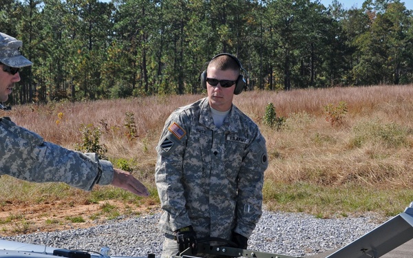 Three U.S. Army soldiers in ACU (Army Combat Uniform) camouflage are preparing or inspecting a small unmanned aerial vehicle (UAV) marked with the number '2378'. The aircraft appears to be mounted on a ground support/launch stand on a gravel surface. The setting appears to be a military training or operational area with a tree line visible in the background.
