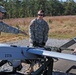 Three U.S. Army soldiers in ACU (Army Combat Uniform) camouflage are preparing or inspecting a small unmanned aerial vehicle (UAV) marked with the number '2378'. The aircraft appears to be mounted on a ground support/launch stand on a gravel surface. The setting appears to be a military training or operational area with a tree line visible in the background.