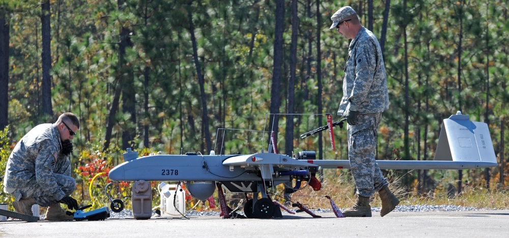 Two U.S. Army soldiers in ACU (Army Combat Uniform) are conducting pre-flight or maintenance checks on a fixed-wing unmanned aerial vehicle (UAV) on a paved surface. The aircraft bears the number '2378' and appears to be an RQ-7 Shadow or similar tactical UAV. The setting appears to be a military airfield or training area surrounded by pine trees.