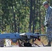 Two U.S. Army soldiers in ACU (Army Combat Uniform) are conducting pre-flight or maintenance checks on a fixed-wing unmanned aerial vehicle (UAV) on a paved surface. The aircraft bears the number '2378' and appears to be an RQ-7 Shadow or similar tactical UAV. The setting appears to be a military airfield or training area surrounded by pine trees.