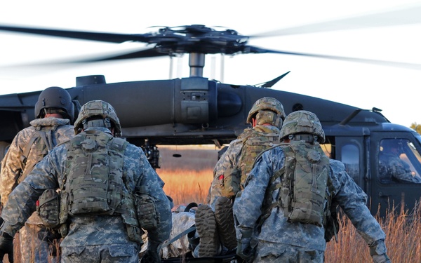 Four soldiers in U.S. Army combat uniforms are carrying a casualty on a litter (stretcher) toward a UH-60 Black Hawk helicopter landed in an open field. The scene depicts a medical evacuation (MEDEVAC) operation, likely during a training exercise given the controlled environment. The image is taken during golden hour lighting in what appears to be a rural or training area with tall dry grass.