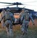 Four soldiers in U.S. Army combat uniforms are carrying a casualty on a litter (stretcher) toward a UH-60 Black Hawk helicopter landed in an open field. The scene depicts a medical evacuation (MEDEVAC) operation, likely during a training exercise given the controlled environment. The image is taken during golden hour lighting in what appears to be a rural or training area with tall dry grass.