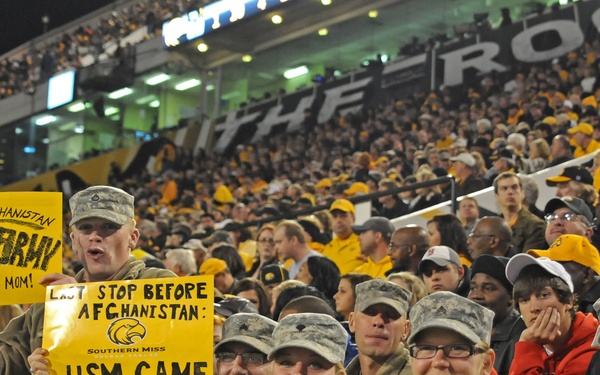 A group of U.S. Army soldiers in camouflage uniforms are attending a Southern Miss (University of Southern Mississippi) football game, holding handmade yellow signs indicating they are deploying to Afghanistan. The signs reference the C Co, 237th BSB (Brigade Support Battalion) and include personal messages to family members. The event appears to be a send-off recognition at a packed night game at what appears to be M.M. Roberts Stadium.
