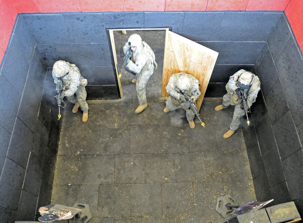 An overhead/bird's-eye view of what appears to be a military close-quarters battle (CQB) training exercise inside a simulated room-clearing facility. Four soldiers in US Army combat uniforms (ACU pattern) are conducting a room entry drill through a plywood doorframe. The scene shows active training with visible smoke/residue and flat cardboard target silhouettes on the floor.