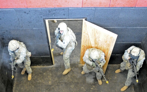 An overhead/bird's-eye view of what appears to be a military close-quarters battle (CQB) training exercise inside a simulated room-clearing facility. Four soldiers in US Army combat uniforms (ACU pattern) are conducting a room entry drill through a plywood doorframe. The scene shows active training with visible smoke/residue and flat cardboard target silhouettes on the floor.