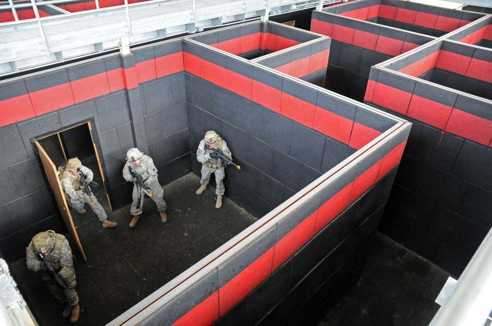 Four military personnel in camouflage uniforms are conducting urban warfare or close-quarters battle (CQB) training in a purpose-built training facility. The structure consists of a maze-like arrangement of rooms with dark gray and red-striped block walls, simulating a building interior. The soldiers are practicing room-clearing techniques, with at least one soldier positioned at a doorway in a tactical stance.