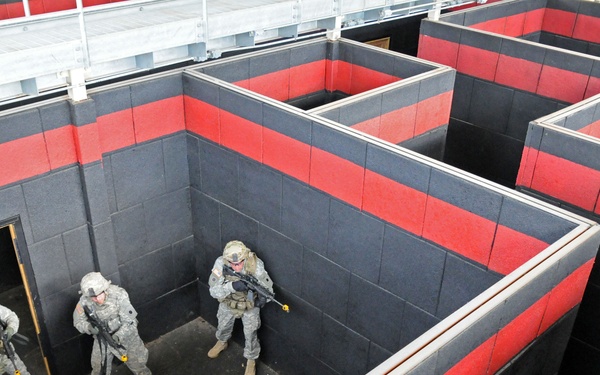 Four military personnel in camouflage uniforms are conducting urban warfare or close-quarters battle (CQB) training in a purpose-built training facility. The structure consists of a maze-like arrangement of rooms with dark gray and red-striped block walls, simulating a building interior. The soldiers are practicing room-clearing techniques, with at least one soldier positioned at a doorway in a tactical stance.