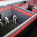 Four military personnel in camouflage uniforms are conducting urban warfare or close-quarters battle (CQB) training in a purpose-built training facility. The structure consists of a maze-like arrangement of rooms with dark gray and red-striped block walls, simulating a building interior. The soldiers are practicing room-clearing techniques, with at least one soldier positioned at a doorway in a tactical stance.