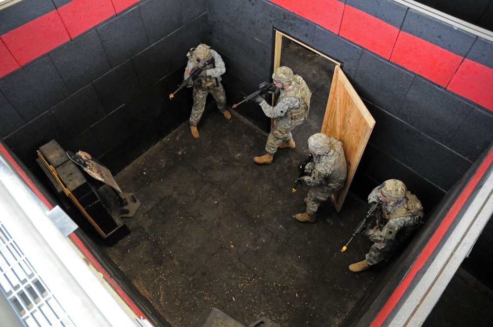 An overhead view shows four military personnel in full combat gear conducting a room-clearing drill inside what appears to be a training facility. The soldiers are wearing ACU (Army Combat Uniform) camouflage and carrying rifles, positioned around a simulated doorway constructed from plywood. The environment features padded black and red walls consistent with an indoor military training range.