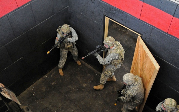 An overhead view shows four military personnel in full combat gear conducting a room-clearing drill inside what appears to be a training facility. The soldiers are wearing ACU (Army Combat Uniform) camouflage and carrying rifles, positioned around a simulated doorway constructed from plywood. The environment features padded black and red walls consistent with an indoor military training range.