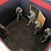 An overhead view shows four military personnel in full combat gear conducting a room-clearing drill inside what appears to be a training facility. The soldiers are wearing ACU (Army Combat Uniform) camouflage and carrying rifles, positioned around a simulated doorway constructed from plywood. The environment features padded black and red walls consistent with an indoor military training range.