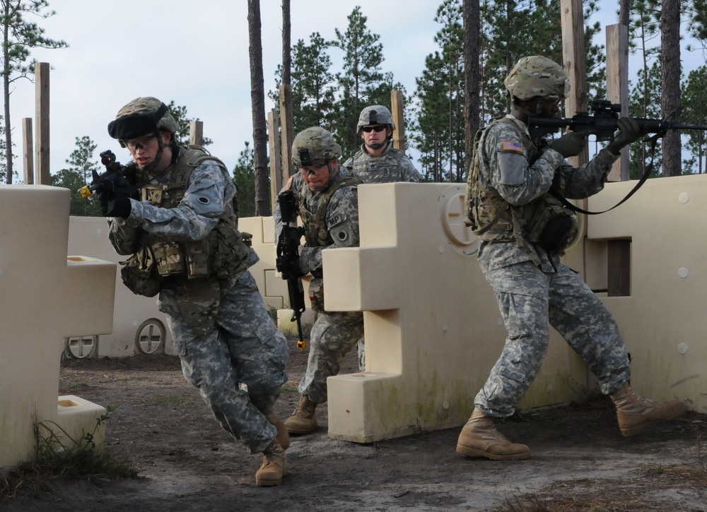 Four U.S. Army soldiers in ACU (Army Combat Uniform) pattern camouflage are conducting a military training exercise at what appears to be a mock urban combat training facility. The soldiers are moving through a simulated building environment constructed from modular plastic barriers, with weapons raised in tactical positions. One soldier has an 'MP' (Military Police) patch visible, and U.S. flag patches are visible on uniforms.