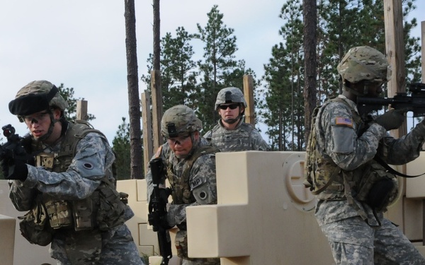 Four U.S. Army soldiers in ACU (Army Combat Uniform) pattern camouflage are conducting a military training exercise at what appears to be a mock urban combat training facility. The soldiers are moving through a simulated building environment constructed from modular plastic barriers, with weapons raised in tactical positions. One soldier has an 'MP' (Military Police) patch visible, and U.S. flag patches are visible on uniforms.