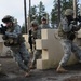 Four U.S. Army soldiers in ACU (Army Combat Uniform) pattern camouflage are conducting a military training exercise at what appears to be a mock urban combat training facility. The soldiers are moving through a simulated building environment constructed from modular plastic barriers, with weapons raised in tactical positions. One soldier has an 'MP' (Military Police) patch visible, and U.S. flag patches are visible on uniforms.