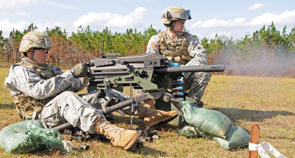 Two U.S. Army soldiers in ACU (Army Combat Uniform) pattern camouflage are conducting a live-fire training exercise on an open range. One soldier is operating what appears to be an M2 .50 caliber heavy machine gun or Mk 19 grenade launcher mounted on a tripod, with visible smoke indicating recent discharge. Sandbags and ammunition are positioned around the weapon system.