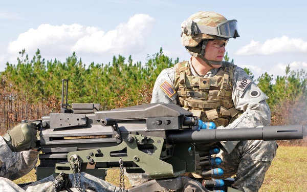 Two U.S. Army soldiers in ACU (Army Combat Uniform) pattern camouflage are conducting a live-fire training exercise on an open range. One soldier is operating what appears to be an M2 .50 caliber heavy machine gun or Mk 19 grenade launcher mounted on a tripod, with visible smoke indicating recent discharge. Sandbags and ammunition are positioned around the weapon system.