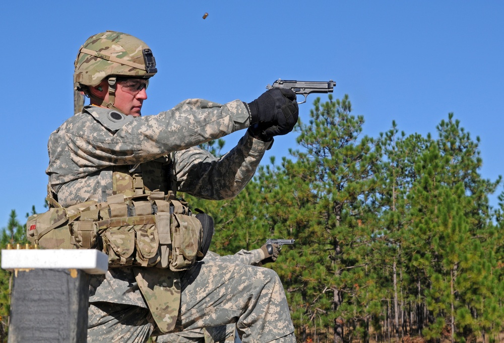A U.S. Army soldier in full combat gear is firing a semi-automatic pistol at what appears to be a military shooting range. A spent brass casing is visibly ejecting from the firearm, captured mid-air above the weapon. A second soldier partially visible in the lower right also appears to be holding a pistol.