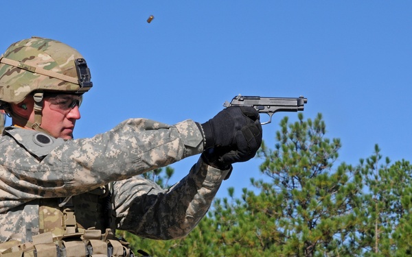 A U.S. Army soldier in full combat gear is firing a semi-automatic pistol at what appears to be a military shooting range. A spent brass casing is visibly ejecting from the firearm, captured mid-air above the weapon. A second soldier partially visible in the lower right also appears to be holding a pistol.