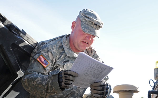 A U.S. Army soldier, identified by name tape as 'GIERE', is conducting a pre-operational vehicle inspection on a military HMMWV (Humvee), referencing a printed checklist document. The soldier is wearing Army Combat Uniform (ACU) with digital camouflage pattern, tactical gloves, and a patrol cap with a U.S. flag patch visible on the left shoulder. Additional soldiers and military vehicles are visible in the background.