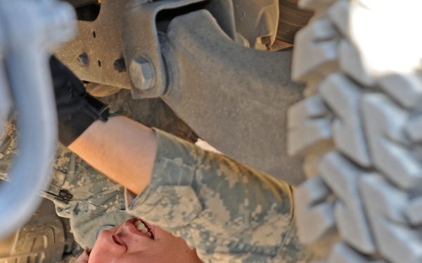 A soldier wearing US Army ACU (Army Combat Uniform) digital camouflage is lying on the ground beneath a military vehicle, performing an inspection or maintenance task. The photo is taken from a low angle looking up, showing the undercarriage of the vehicle and a large off-road tire in the foreground. The soldier appears to be examining or working on a component of the vehicle's underside.