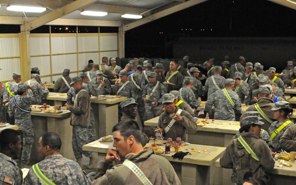 A large group of military personnel in U.S. Army combat uniforms are gathered in what appears to be a field dining facility (DFAC) or mess hall. Soldiers are eating meals at concrete bench-style tables, with small American flags visible as decorations suggesting a holiday meal, possibly Thanksgiving. The facility appears to be a semi-permanent structure with corrugated metal walls and overhead lighting.