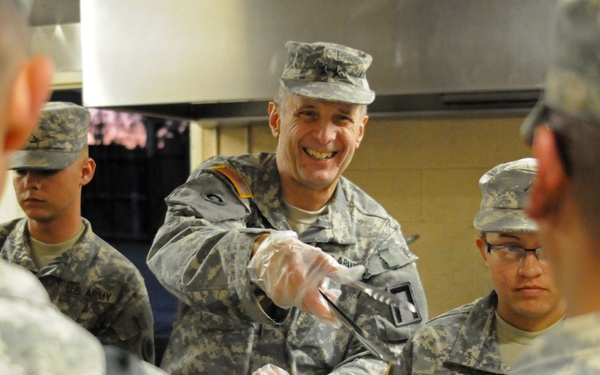 A group of U.S. Army soldiers are in a military dining facility (DFAC). A smiling senior soldier wearing latex gloves is serving food to other soldiers in a chow line. The atmosphere appears positive and informal, with the server engaging cheerfully with those being served.