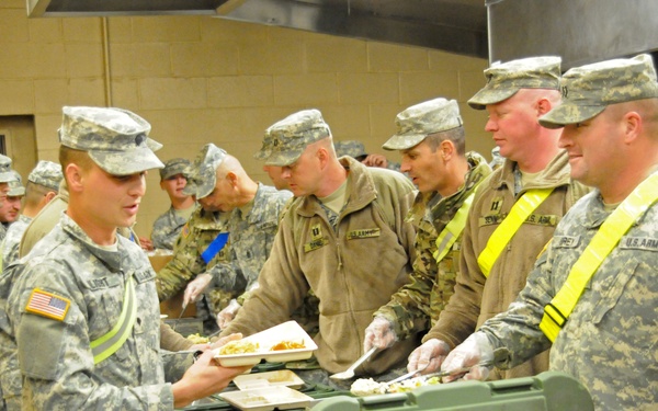 U.S. Army soldiers are going through a military field kitchen chow line, being served food by fellow soldiers wearing gloves. The setting appears to be an indoor facility, likely a dining facility or field kitchen setup with insulated food transport containers. Several soldiers are wearing high-visibility yellow reflective vests over their ACU (Army Combat Uniform) camouflage.