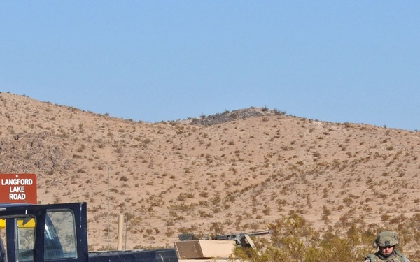 A military training exercise or checkpoint operation in a desert environment, likely in the American Southwest based on the terrain and road sign. Soldiers in U.S. Army combat uniforms are interacting with a civilian dark blue pickup truck with its hood open, alongside military HMMWVs (Humvees). The scene appears to depict a vehicle stop or inspection scenario.