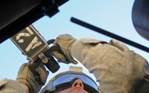 A military service member in full combat gear is photographed from a low angle, appearing to adjust or inspect equipment mounted above them, likely on a vehicle or aircraft. The soldier is wearing a tactical helmet with a mounted device, ballistic goggles, multicam/OCP pattern uniform, tactical vest, and gloves. The perspective is an upward-looking shot that captures the blue sky in the background.