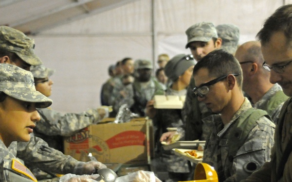 Military personnel are gathered in what appears to be a field dining facility or mess tent, where soldiers in U.S. Army ACU (Army Combat Uniform) camouflage are serving food to fellow service members in a cafeteria-style line. A female soldier wearing plastic gloves is actively ladling what appears to be a sauce or gravy onto a tray held by a male soldier wearing glasses. The setting suggests a forward operating base or deployed environment meal service.