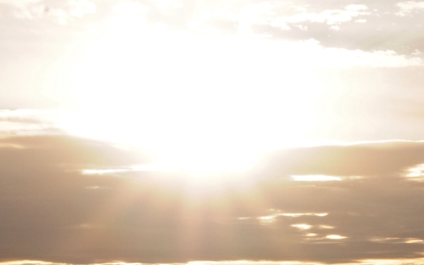 A wide panoramic landscape photograph taken at either sunrise or sunset, with a brilliant sun breaking through clouds over rolling hills or low mountains. A dust cloud or haze hangs low across the flat terrain in the foreground, partially obscuring what appear to be vehicles or machinery silhouetted along the horizon. The warm golden light creates dramatic contrast against the dark landforms and atmospheric dust.