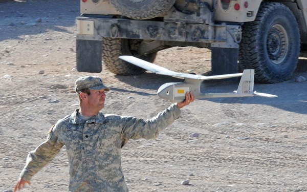 A U.S. Army soldier in ACU (Army Combat Uniform) is hand-launching a small fixed-wing unmanned aerial vehicle (UAV), consistent with an RQ-11 Raven or similar man-portable drone. The soldier is in a wide stance, arm extended, preparing to release the aircraft in an arid, rocky outdoor environment. A military HMMWV (Humvee) is visible in the background.