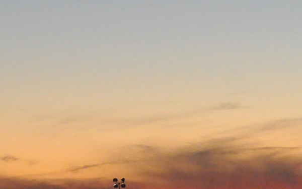 A wide panoramic photograph captured at dusk or dawn showing a dramatic orange and red sky over a silhouetted mountain range. A tall light pole or antenna structure is visible in the middle ground, along with what appears to be a small building or structure on the right. The landscape appears to be an open, flat valley or plain with distant hills.