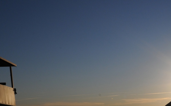 A silhouetted guard or sentry is positioned in a military-style watchtower at sunrise or sunset, overlooking a valley with mountains in the background. Low-lying fog or haze fills the valley floor, while the sun creates a dramatic starburst effect near the horizon. The scene suggests a forward operating base or military outpost in a mountainous region.