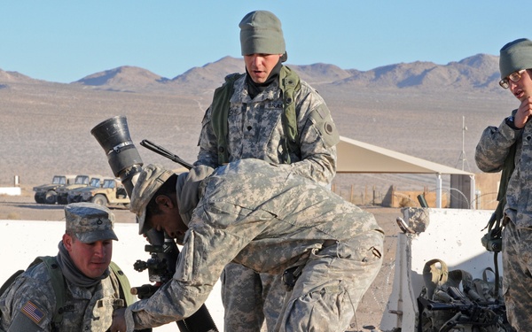 Four U.S. Army soldiers in ACU (Army Combat Uniform) pattern camouflage are working together to set up a mortar system in a desert training environment. The soldiers appear to be conducting a training exercise, with two actively positioning the mortar tube and baseplate while a third observes and a fourth monitors from a standing position. The setting appears to be a military training installation in an arid, desert region.