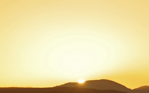 A panoramic landscape photograph capturing a sun setting or rising just behind a silhouetted mountain or hill range. The sky is dominated by warm golden and orange tones radiating from the bright solar disk. A hazy, misty foreground adds atmospheric depth to the scene.