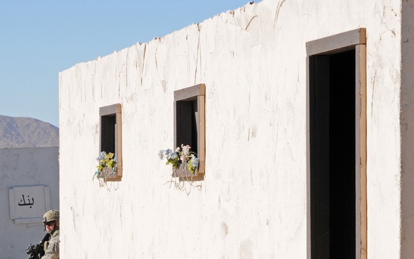 A soldier in full combat gear and camouflage uniform is crouching against a whitewashed building in what appears to be a military training facility or mock village. The scene includes props designed to simulate a civilian environment, including a children's bicycle, flower boxes on windowsills, and Arabic script on a nearby building. The arid, desert landscape with mountains in the background suggests a location consistent with a U.S. military training installation.
