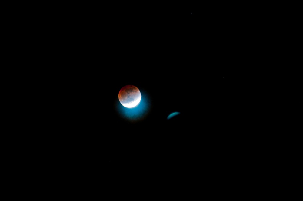 The image shows a lunar eclipse captured against a completely dark night sky. The moon displays a partial eclipse with a reddish-orange (blood moon) coloration on the upper portion and a bright white crescent on the lower left, indicating Earth's shadow partially covering the lunar surface. A smaller, dimmer secondary object — likely an optical artifact, lens flare, or a nearby celestial body — is visible to the right of the moon.