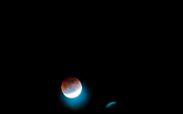 The image shows a lunar eclipse captured against a completely dark night sky. The moon displays a partial eclipse with a reddish-orange (blood moon) coloration on the upper portion and a bright white crescent on the lower left, indicating Earth's shadow partially covering the lunar surface. A smaller, dimmer secondary object — likely an optical artifact, lens flare, or a nearby celestial body — is visible to the right of the moon.