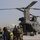 A group of military personnel in combat gear are boarding or preparing to board a CH-47 Chinook tandem-rotor helicopter on what appears to be a forward operating base in an arid environment. The soldiers are equipped with helmets, body armor, and large rucksacks, with one individual wearing a high-visibility yellow marker. The scene suggests a troop insertion, extraction, or air assault operation.