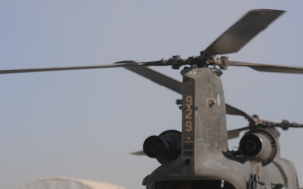 A group of military personnel in combat gear are boarding or preparing to board a CH-47 Chinook tandem-rotor helicopter on what appears to be a forward operating base in an arid environment. The soldiers are equipped with helmets, body armor, and large rucksacks, with one individual wearing a high-visibility yellow marker. The scene suggests a troop insertion, extraction, or air assault operation.